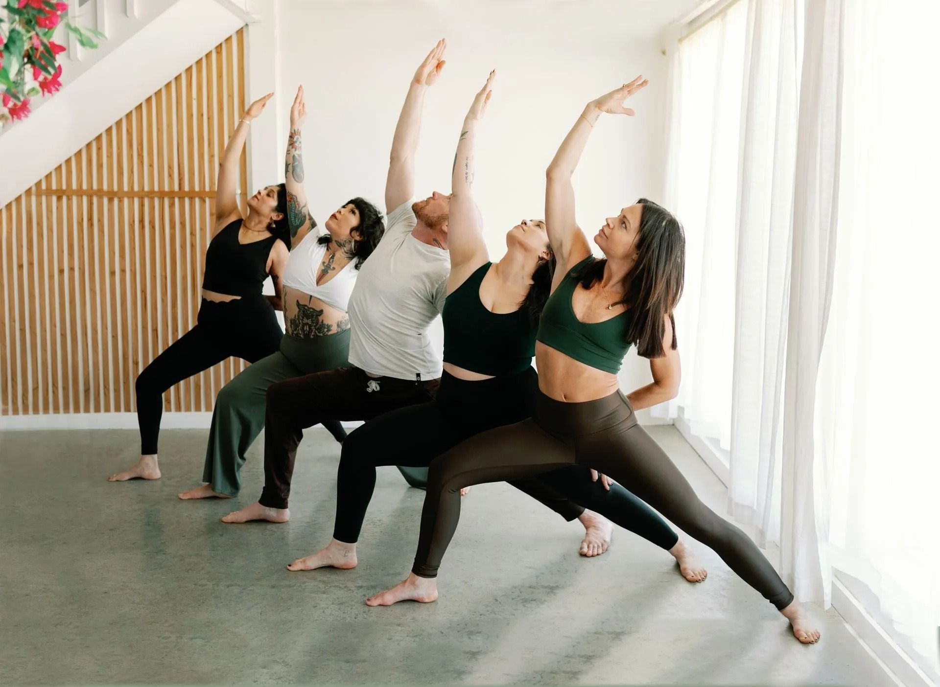 Five people in a bright, modern studio practice yoga in a warrior pose with arms raised against a slatted wood wall.