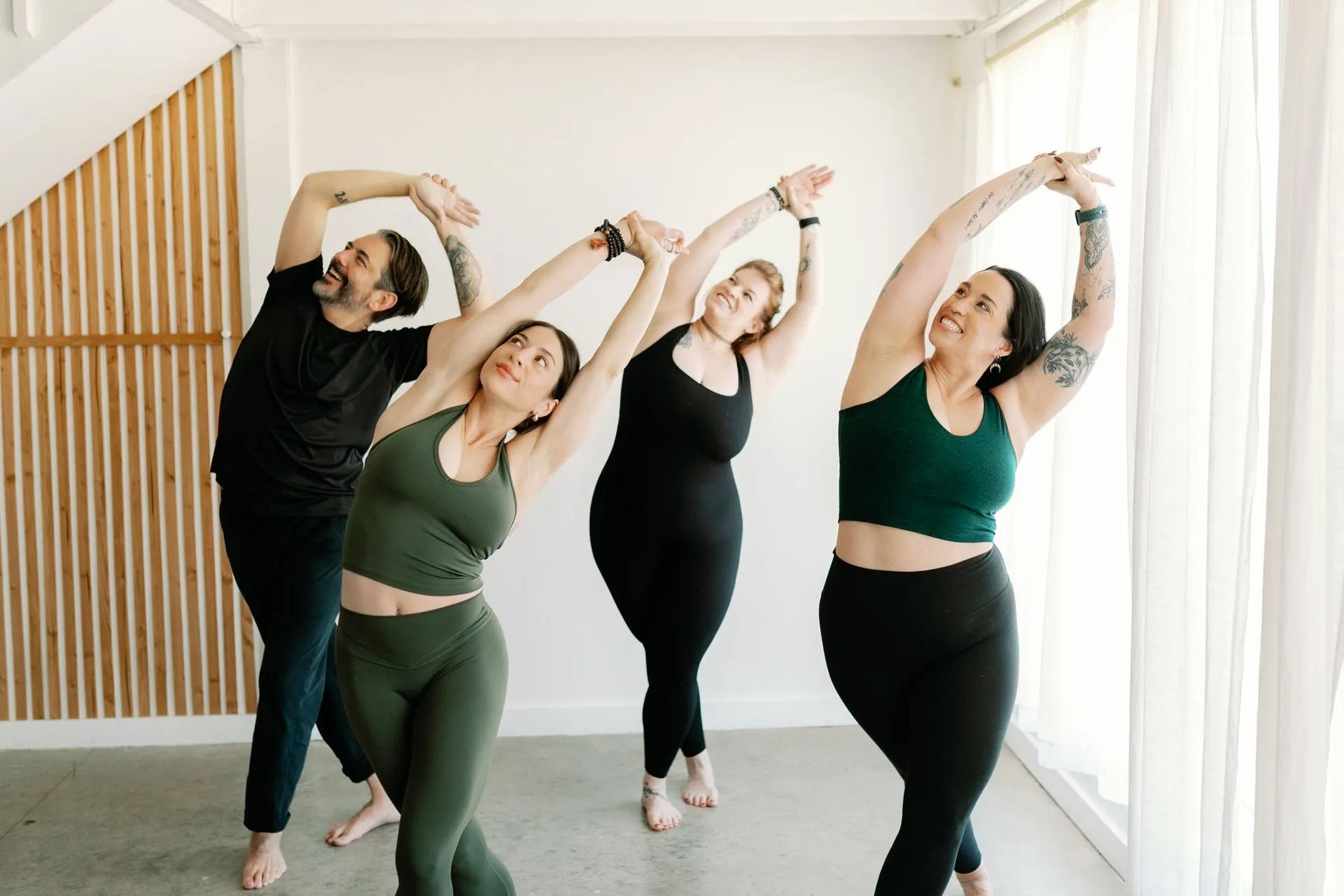Four people in athletic wear pose in a bright studio, smiling while stretching their arms overhead in a side bend.