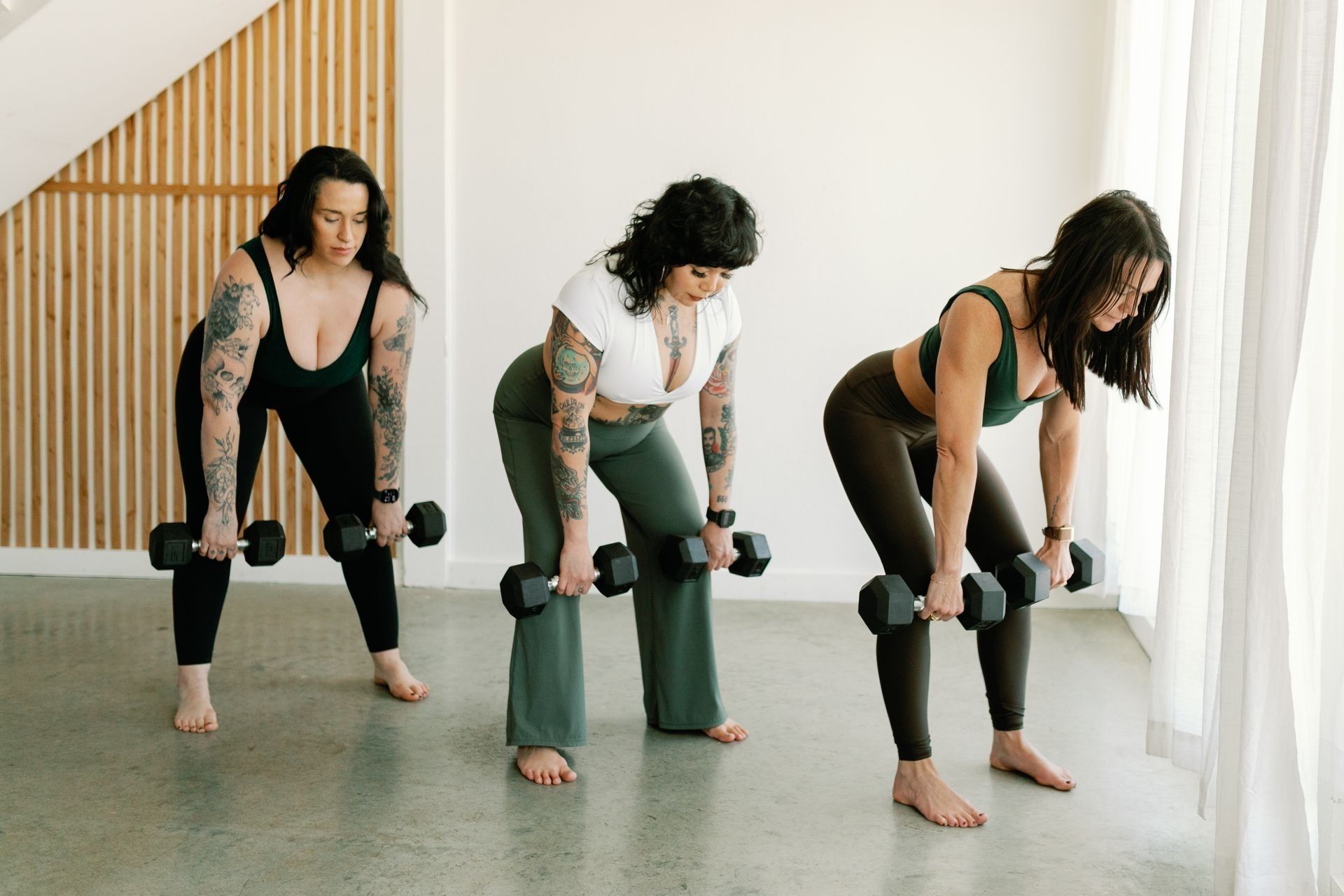 Three people in a bright studio performing a bent-over dumbbell row exercise, wearing athletic clothing.