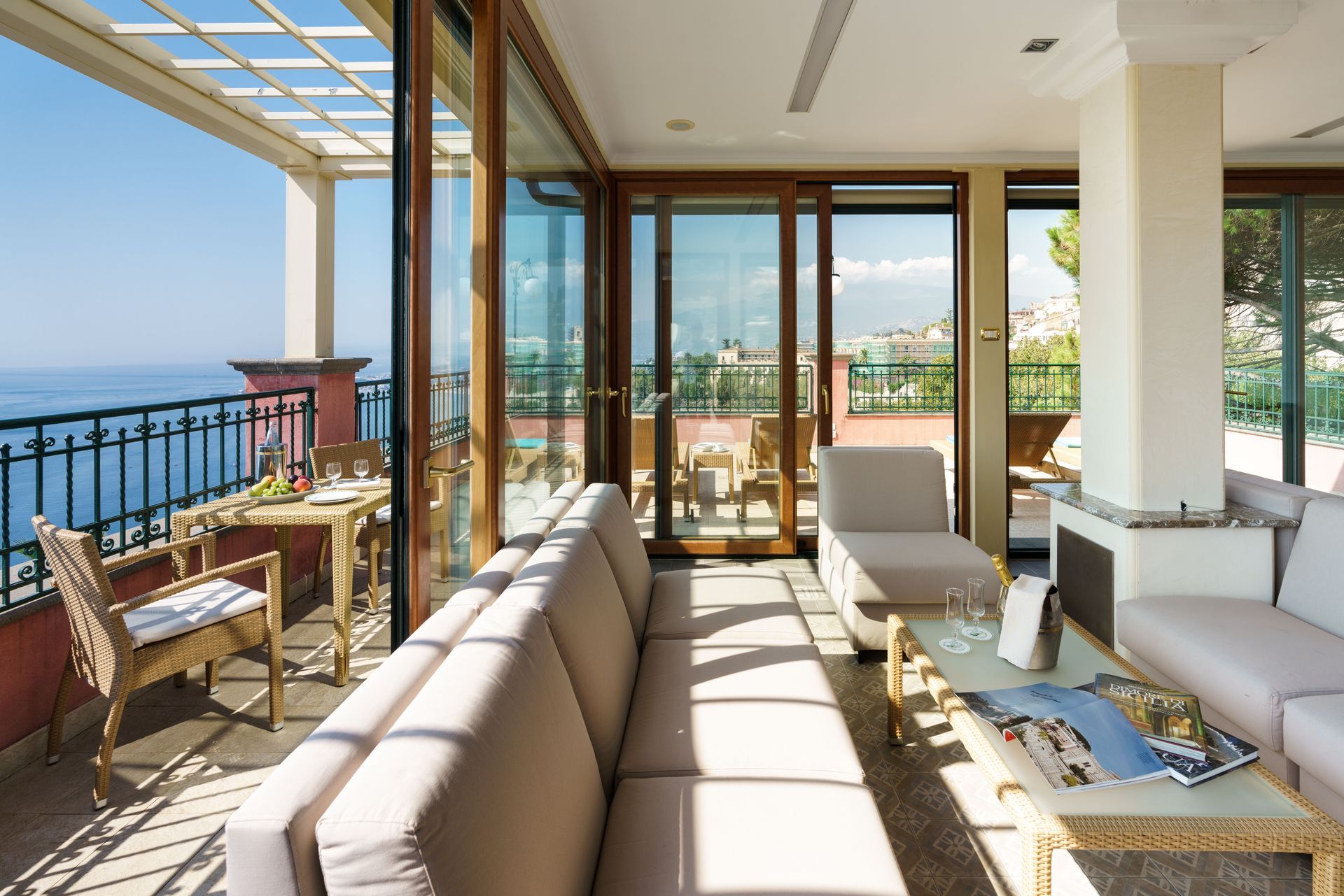 Sunlit luxury patio with sea view, white sofas, glass doors, and wicker chairs overlooking the balcony