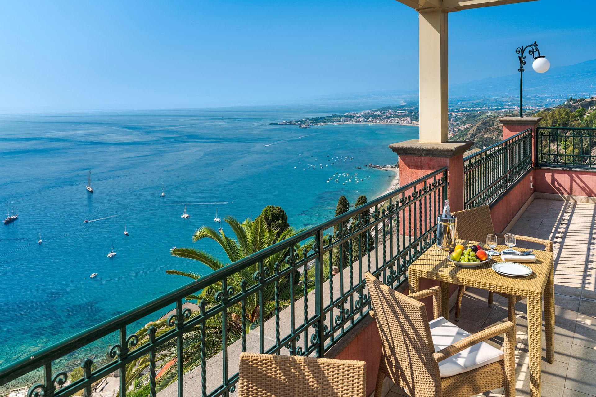 Balcony overlooking a bright blue sea, with chairs, a table, and a coastal view at sunset.