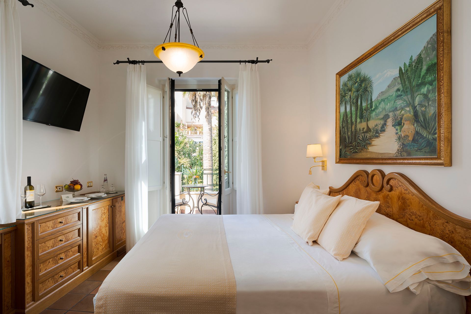 Bright bedroom with a bed, dresser, wall art, TV, and balcony doors opening to greenery