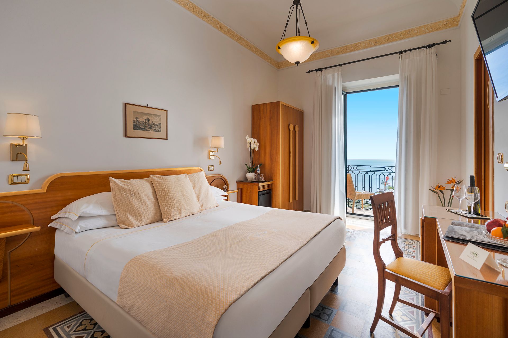 Bright hotel bedroom with a king bed, desk, and balcony doors overlooking the sea.