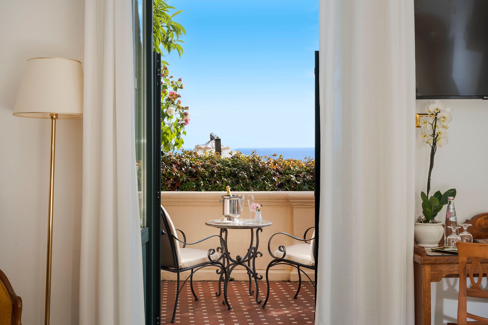 Cozy balcony with wrought-iron table, chairs, white curtains, and ocean view beyond greenery