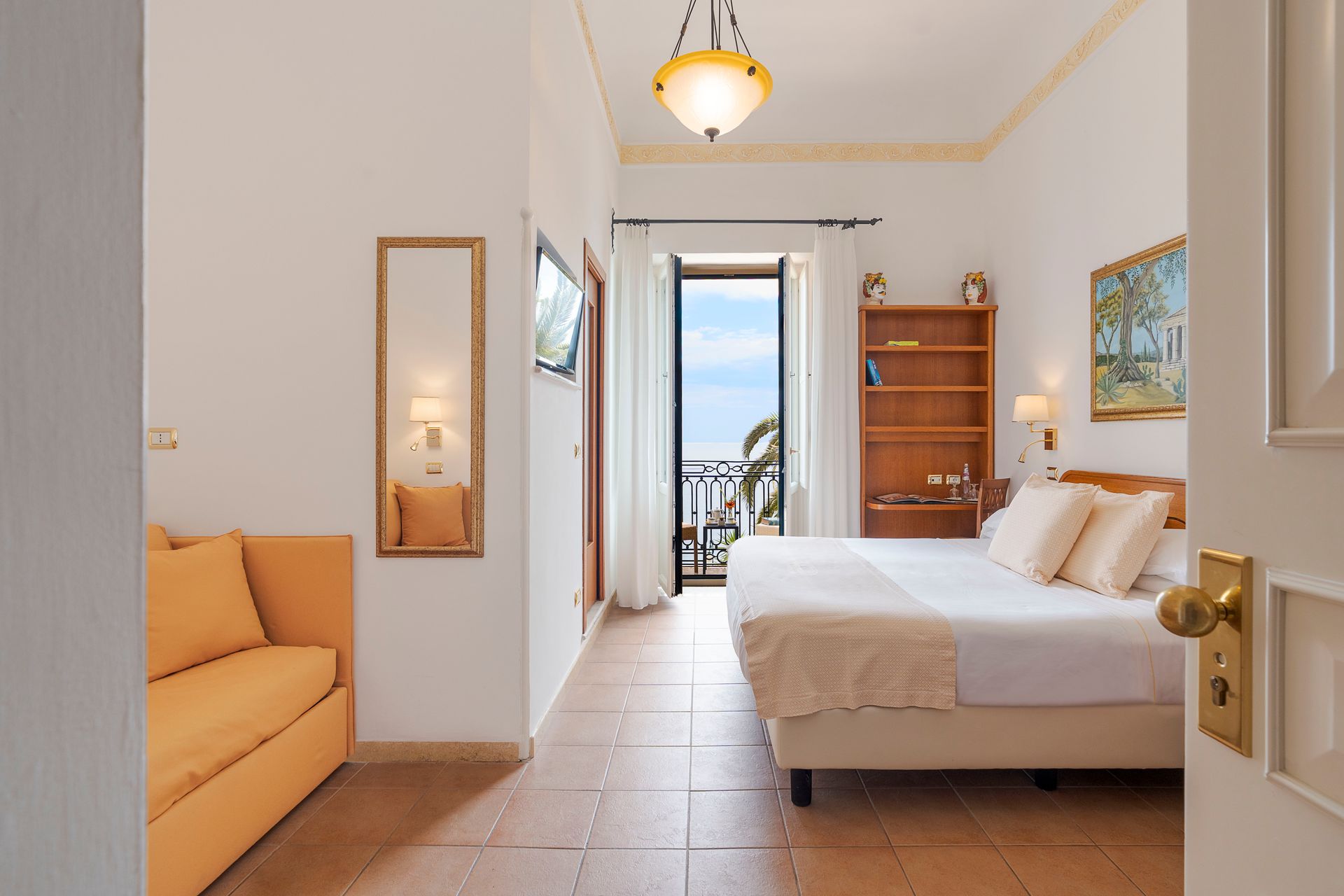 Bright bedroom with a bed, sofa, mirror, bookshelf, and balcony view through open doors