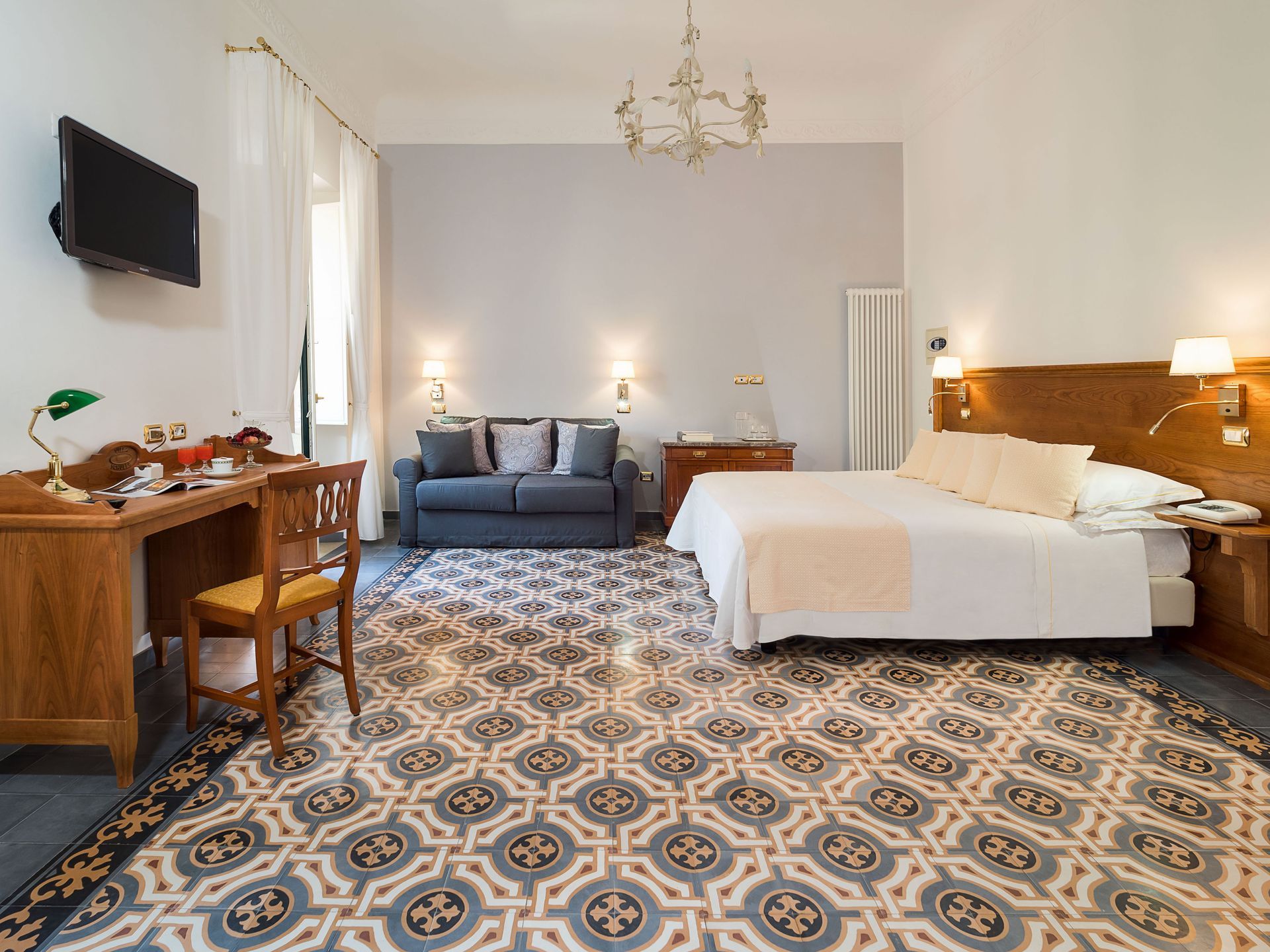 Bright hotel room with a bed, blue sofa, desk, wall TV, and patterned tile floor