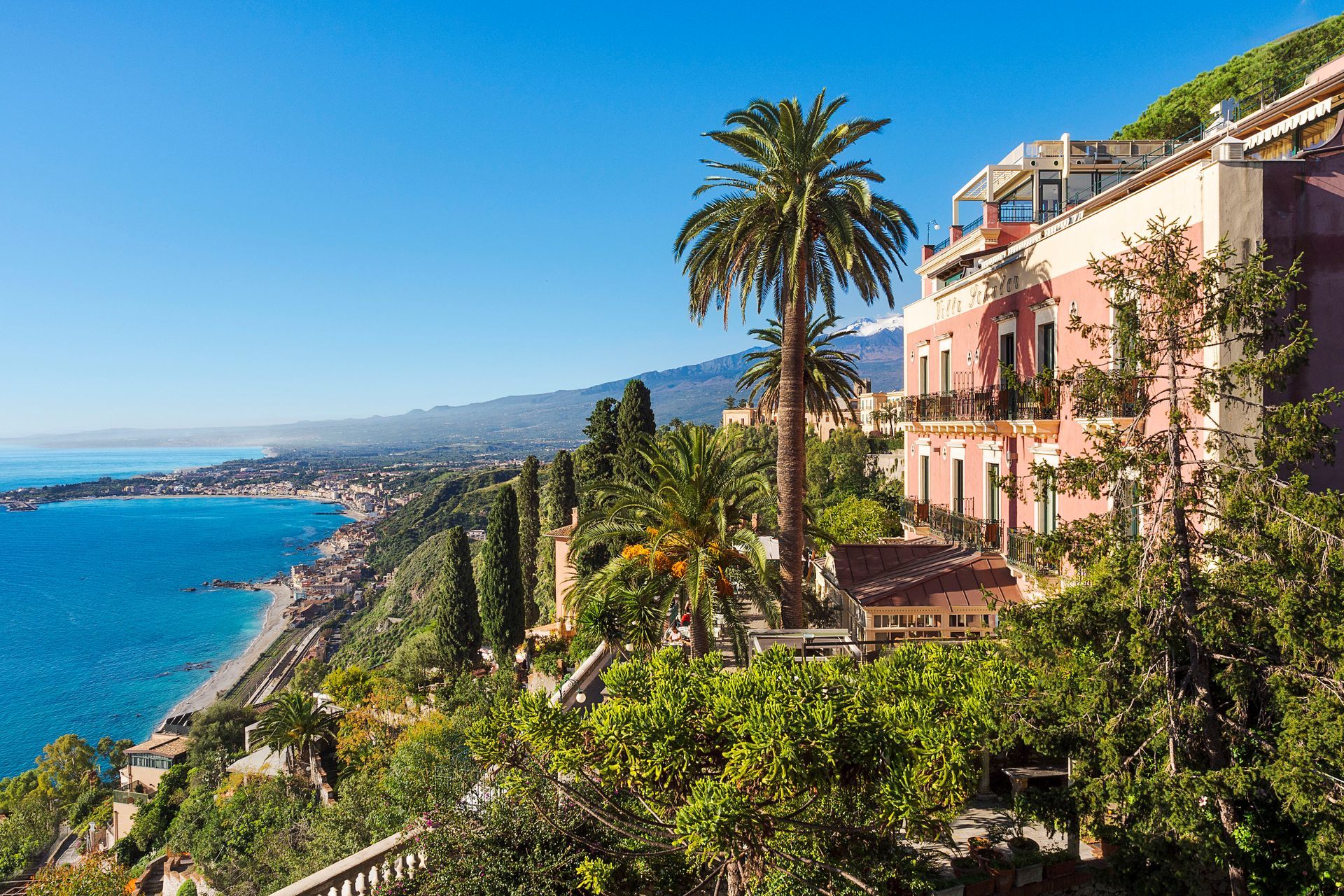 Sunlit seaside hillside with pink villa, palm trees, and blue bay under a clear sky