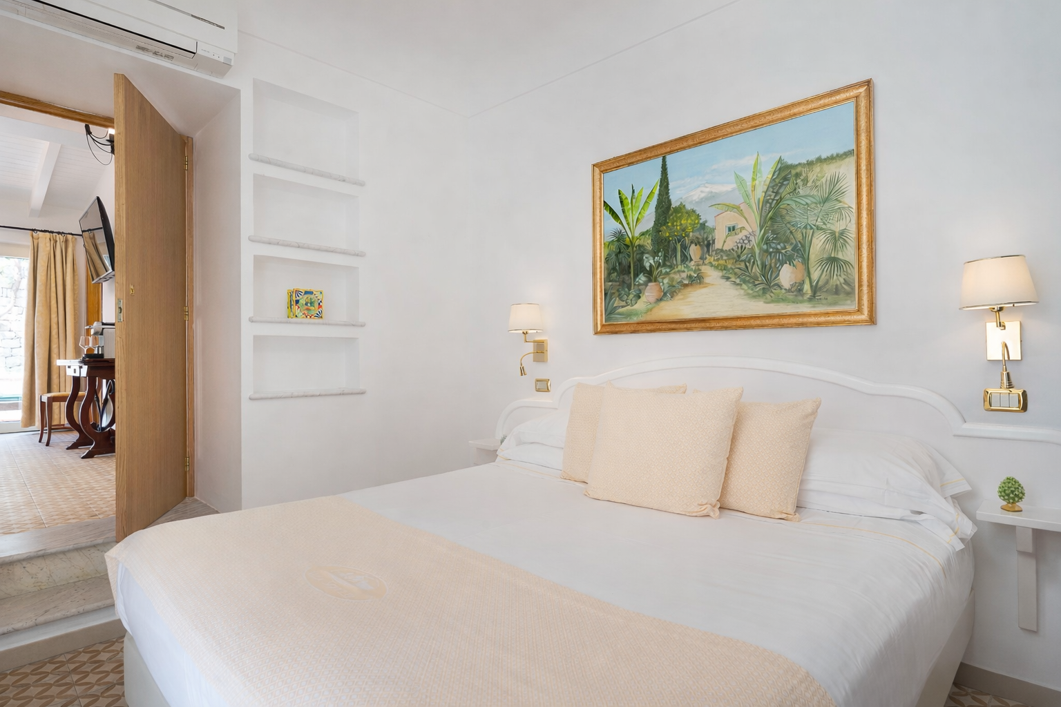 Chambre lumineuse avec un lit blanc, des oreillers beiges, des tableaux et une porte ouverte donnant sur une autre pièce.