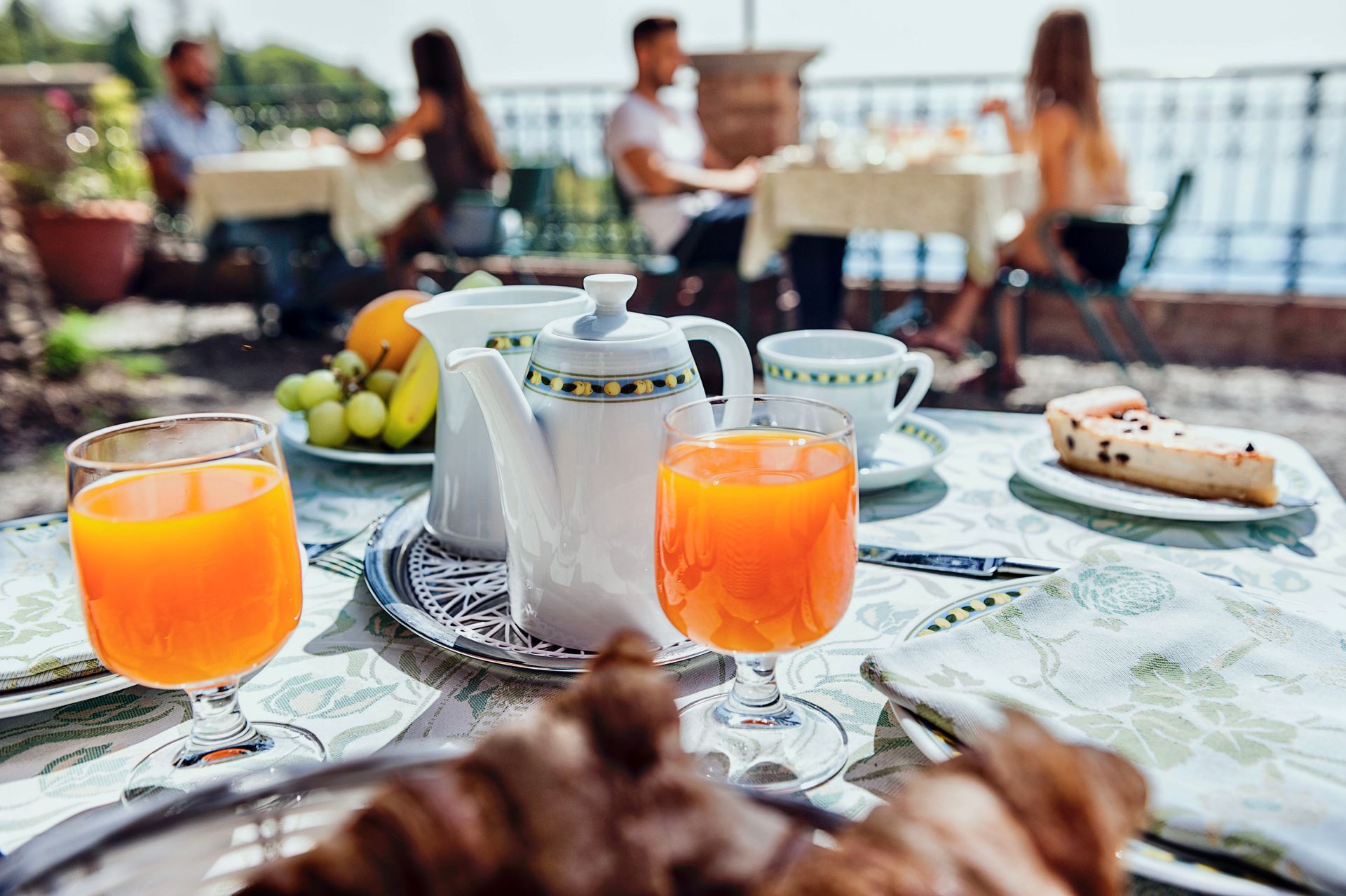 Breakfast table with orange juice, teapot, fruit, and pastries outdoors by the sea