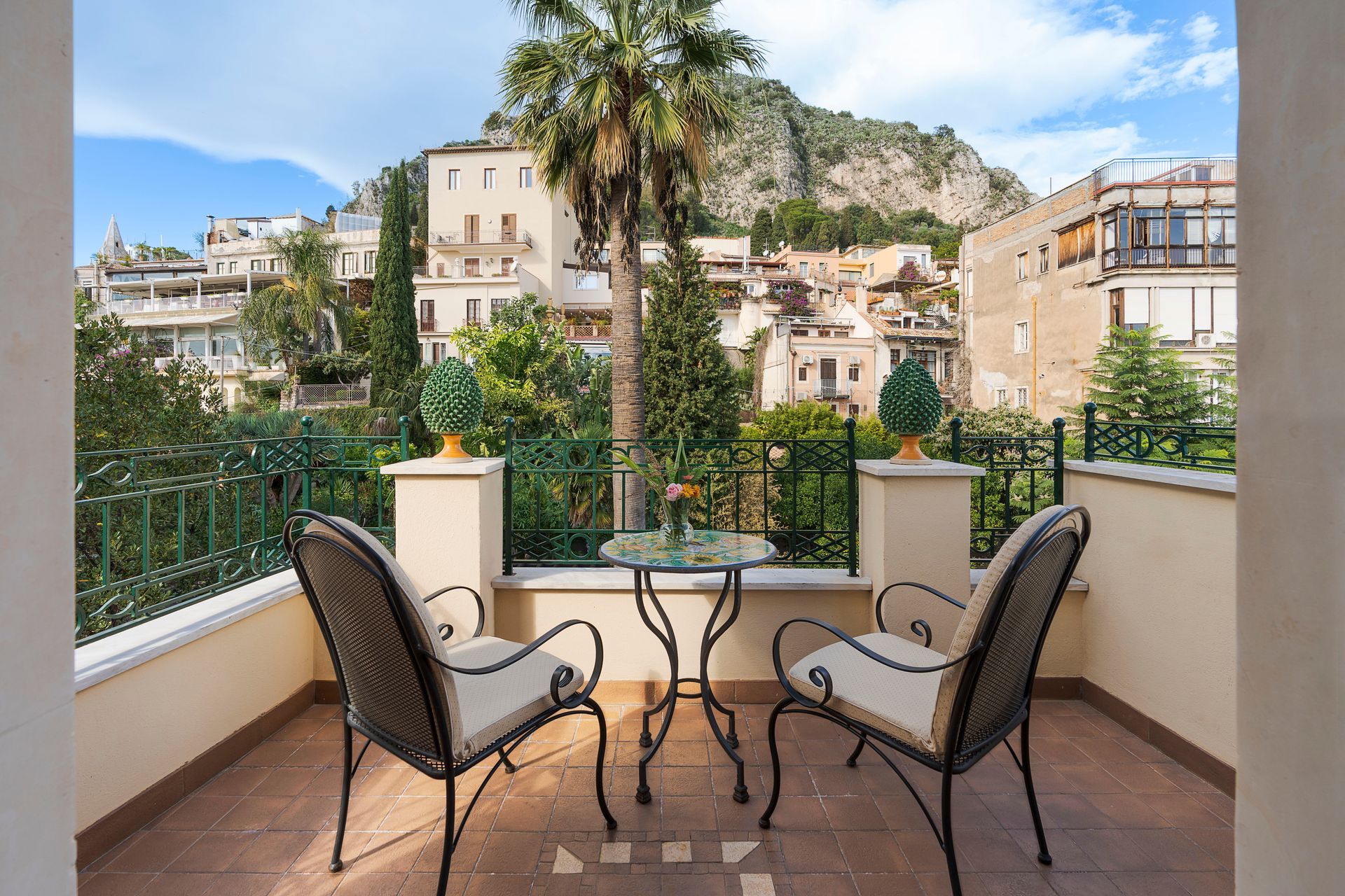 Balcony with two chairs and a table overlooking a sunlit hillside village and palm trees