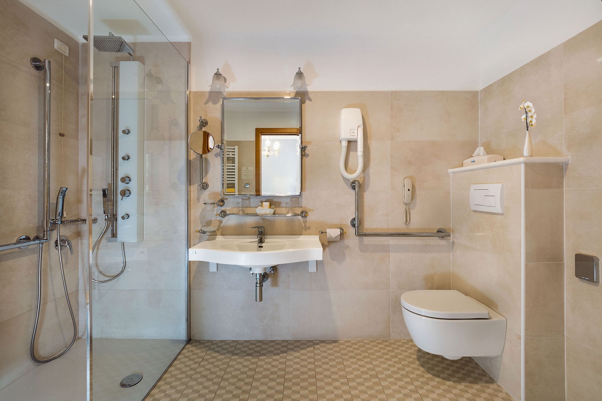 Accessible bathroom with roll-in shower, sink, and wall-mounted toilet in beige tile.