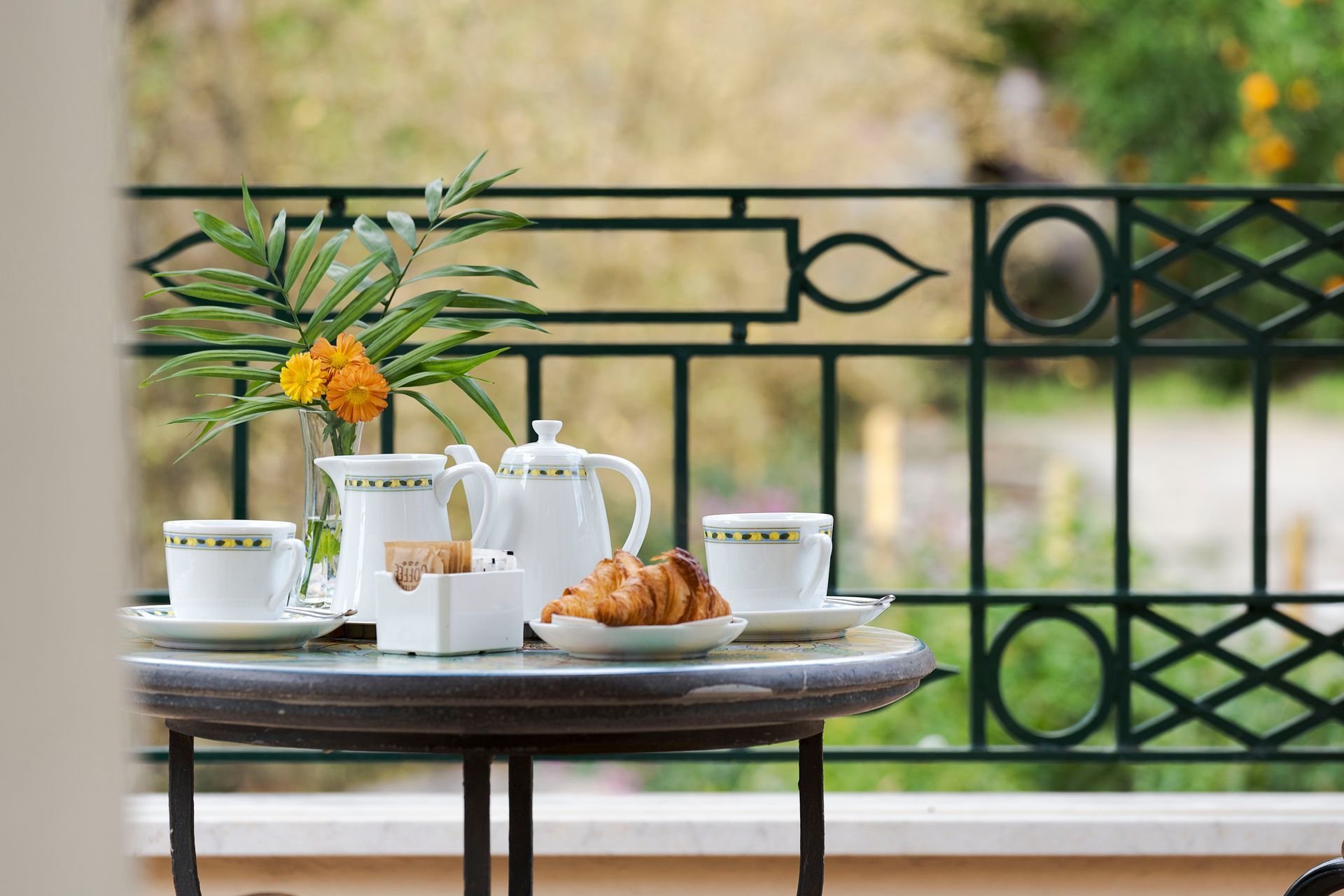 Tea set and pastries on a table by a balcony railing, with flowers in a vase and a garden view.