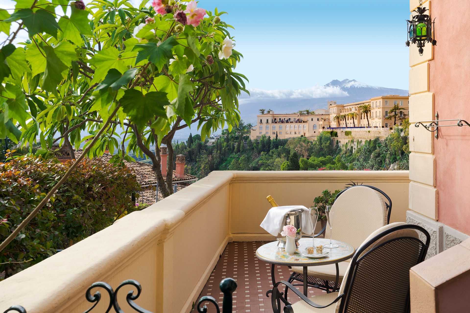 Balcony with patio table and chairs overlooking greenery and a sunlit seaside town.