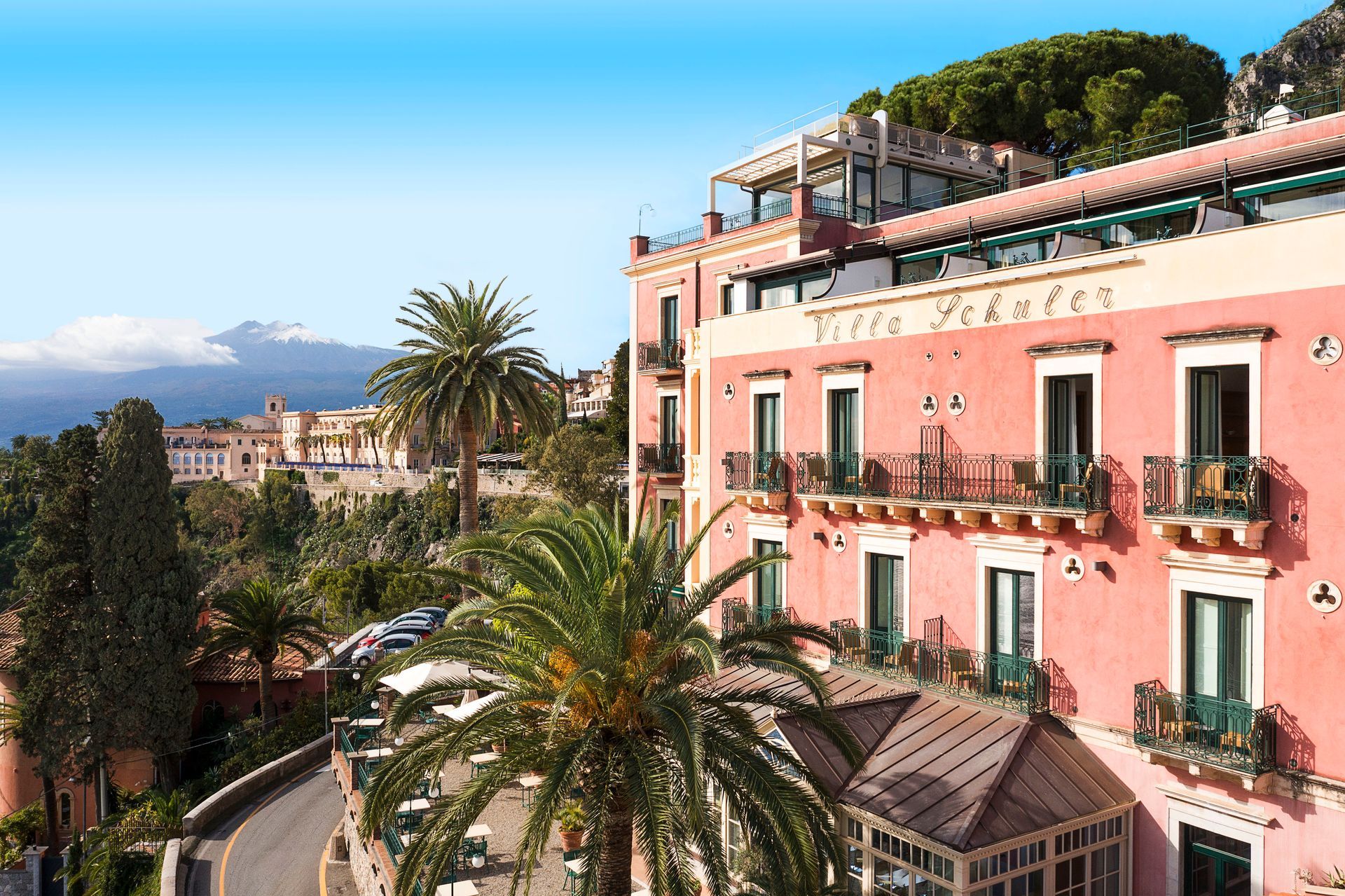 Pink Mediterranean-style buildings with balconies and palm trees on a sunny hillside, sea in the background