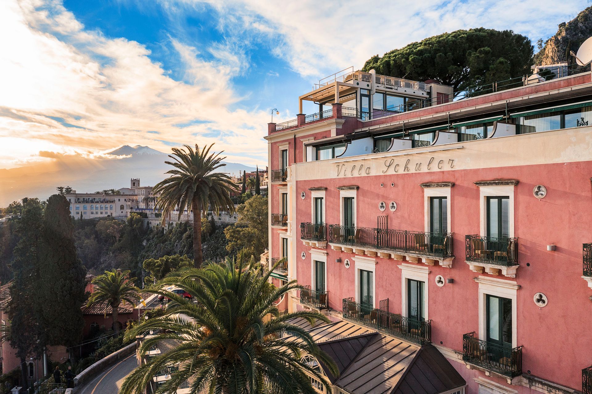 Pink hillside building with balconies and palm trees at sunset under a cloudy sky