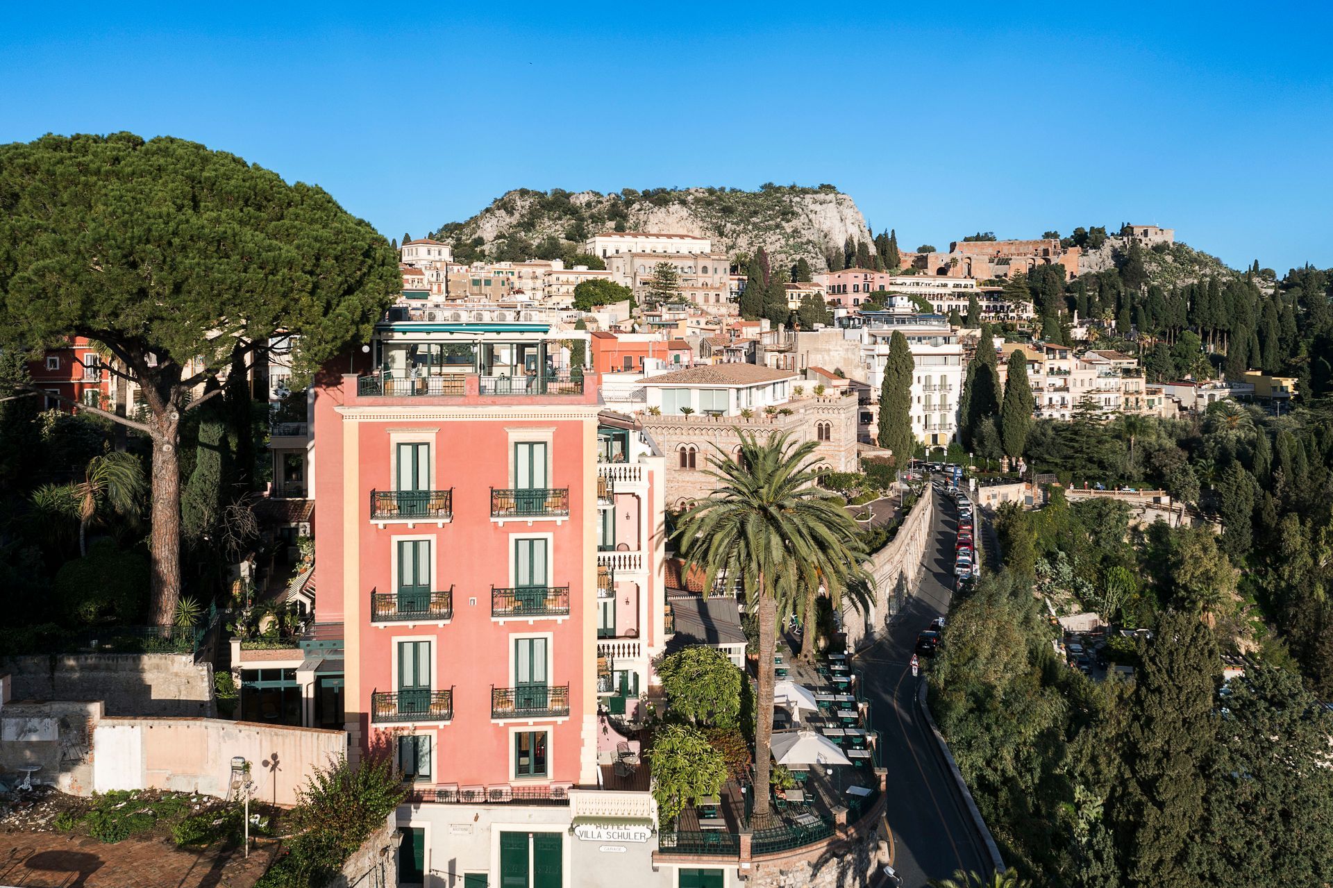 Pink apartment building overlooking a hillside town with trees and rocky cliffs in the background
