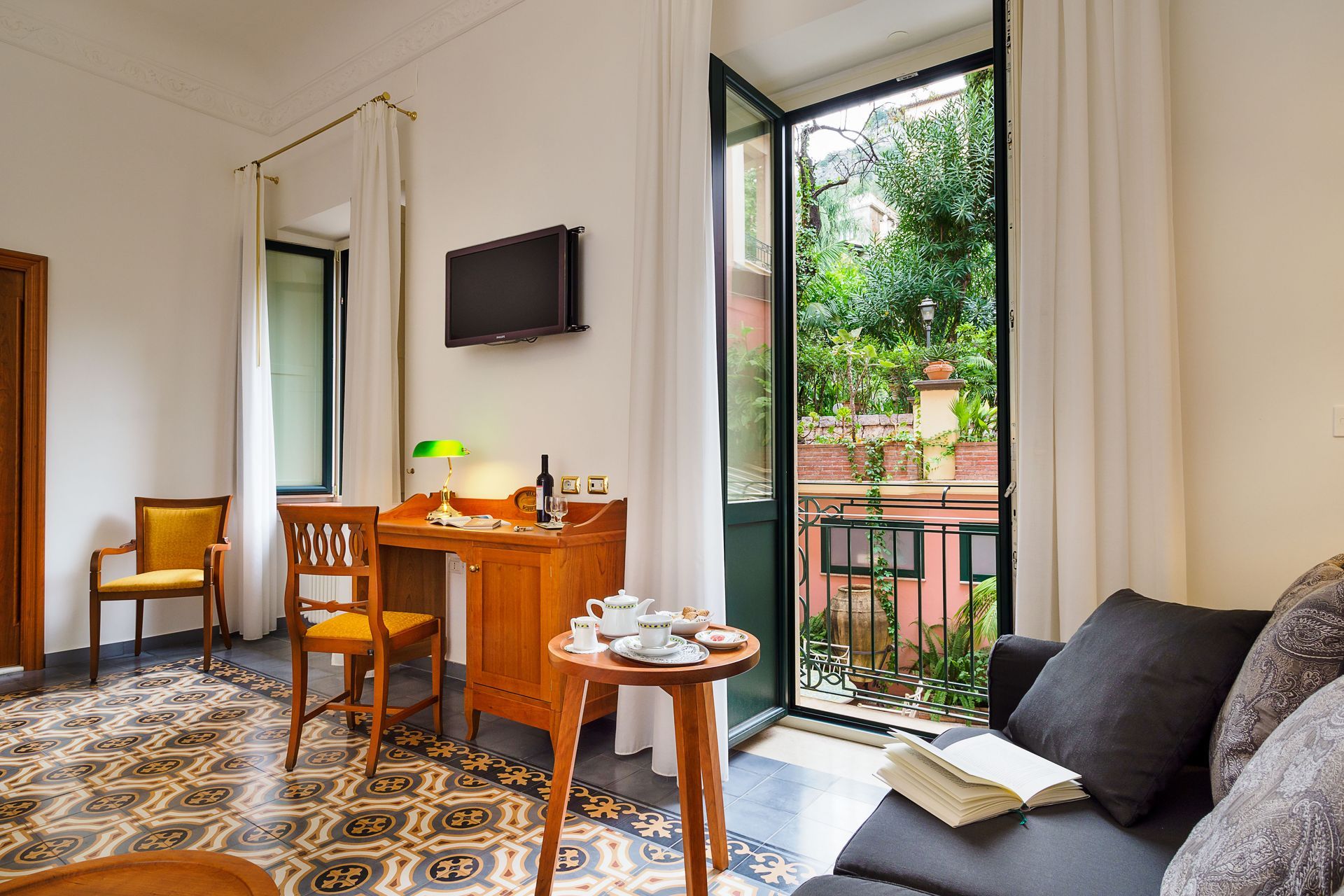 Bright room with desk, wall-mounted TV, sofa, and open balcony door overlooking greenery