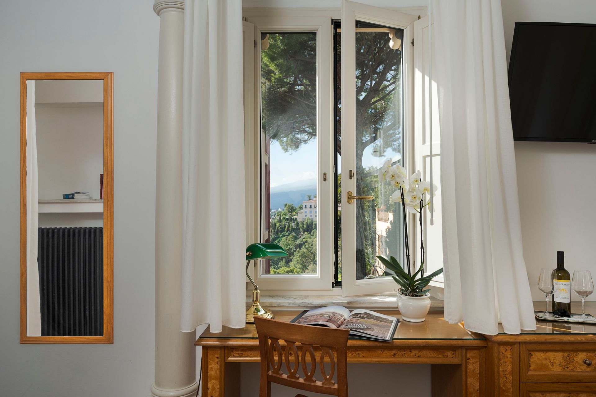 Desk by a bright window with white curtains, potted plants, and a wall mirror in a tidy room.