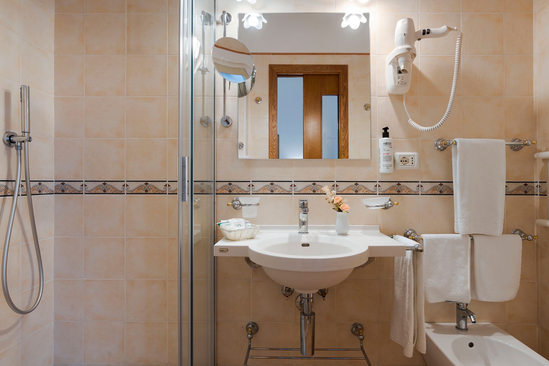 Accessible bathroom with shower, sink, mirror, towel bars, and grab bars in beige tile.