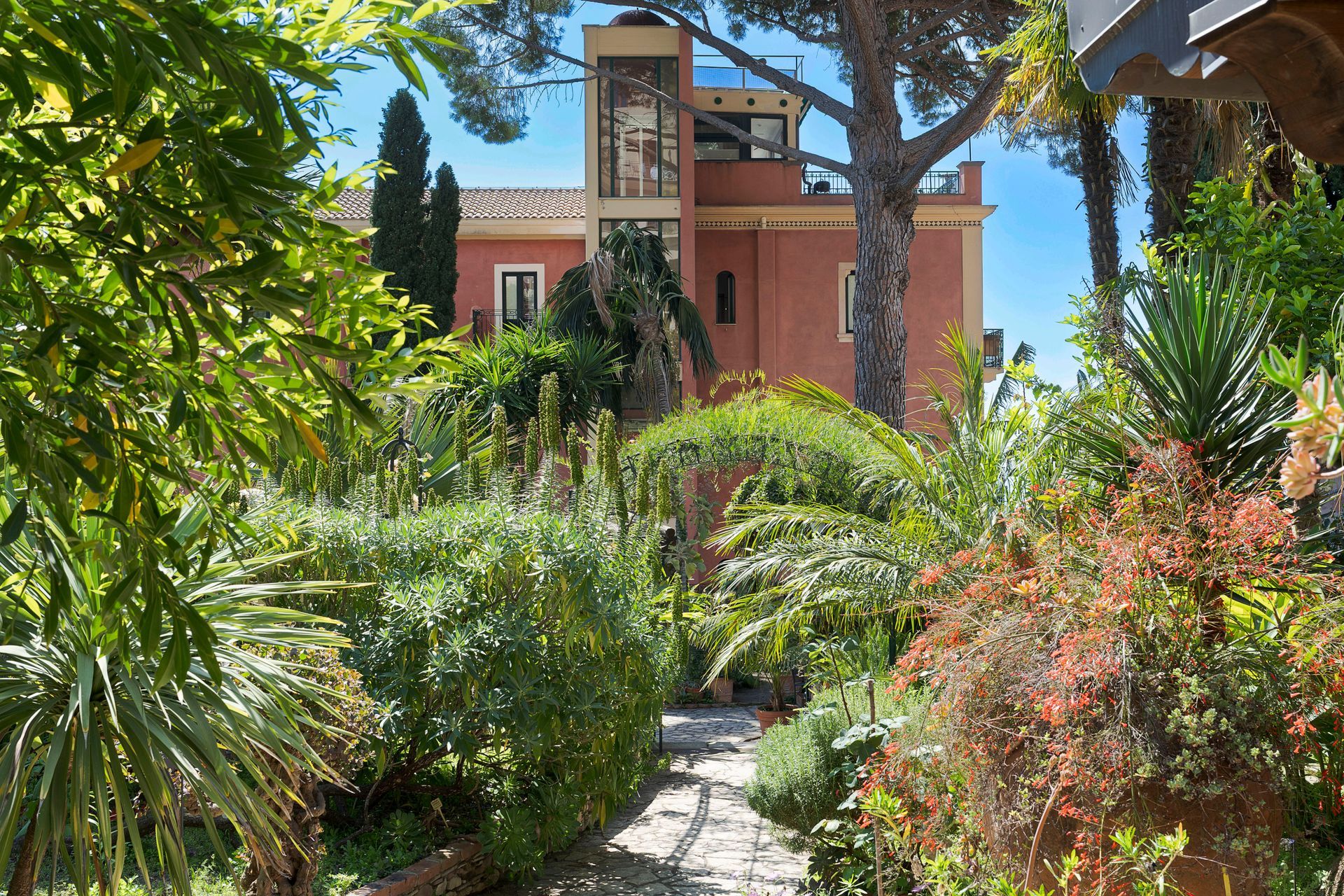 Lush garden path leading to a red-brick house with a wooden balcony and tall trees.
