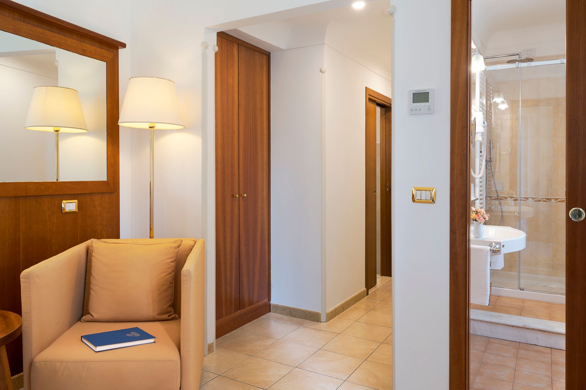 Hotel hallway with beige chair, floor lamp, mirror, and bathroom entrance.