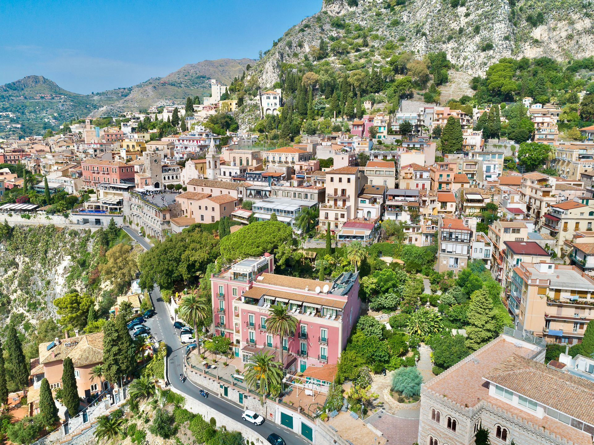 Hillside coastal town with pastel buildings, winding road, and green cliffs under a sunny sky