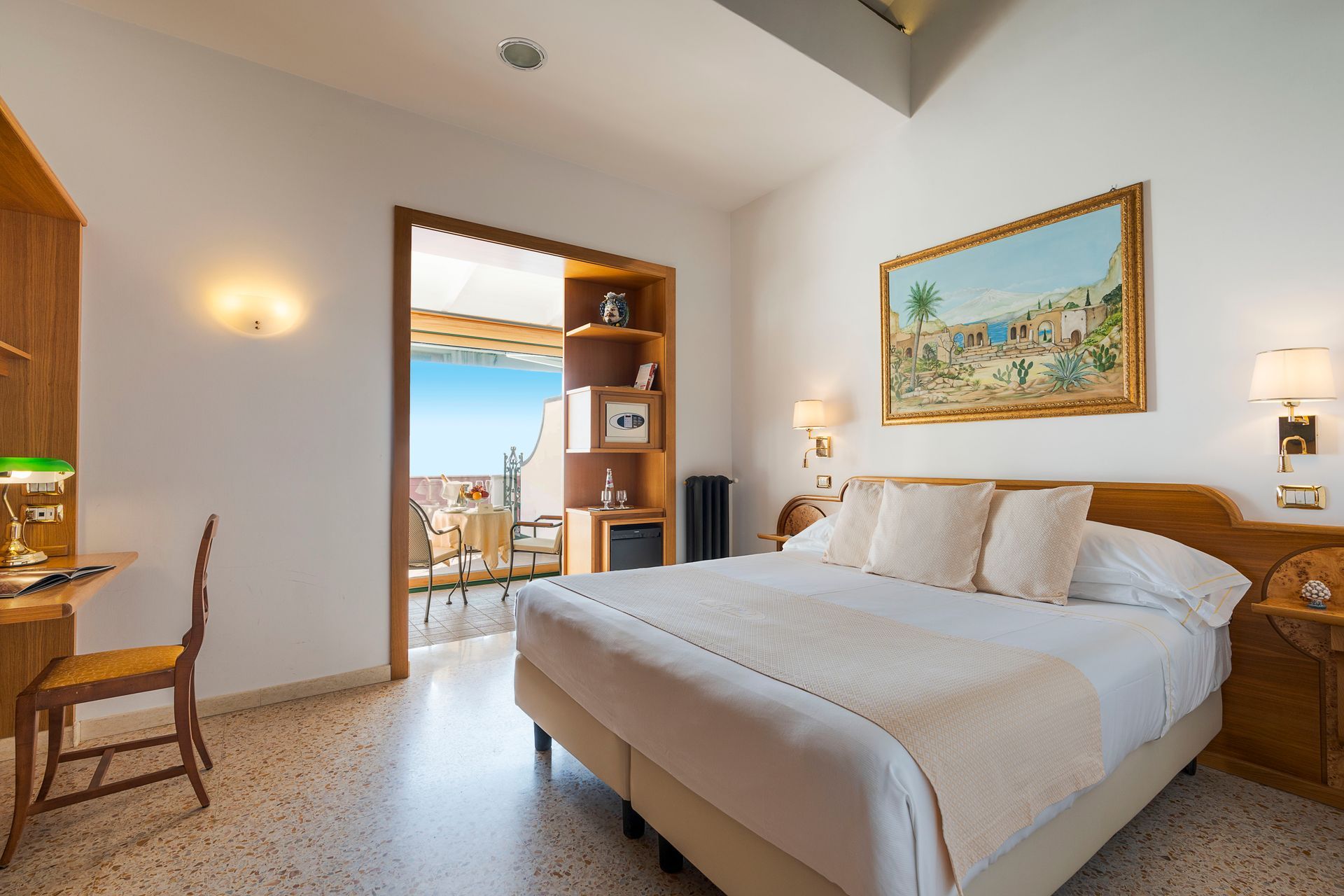 Bright hotel bedroom with a large bed, wall art, lamps, and a balcony view through open doors
