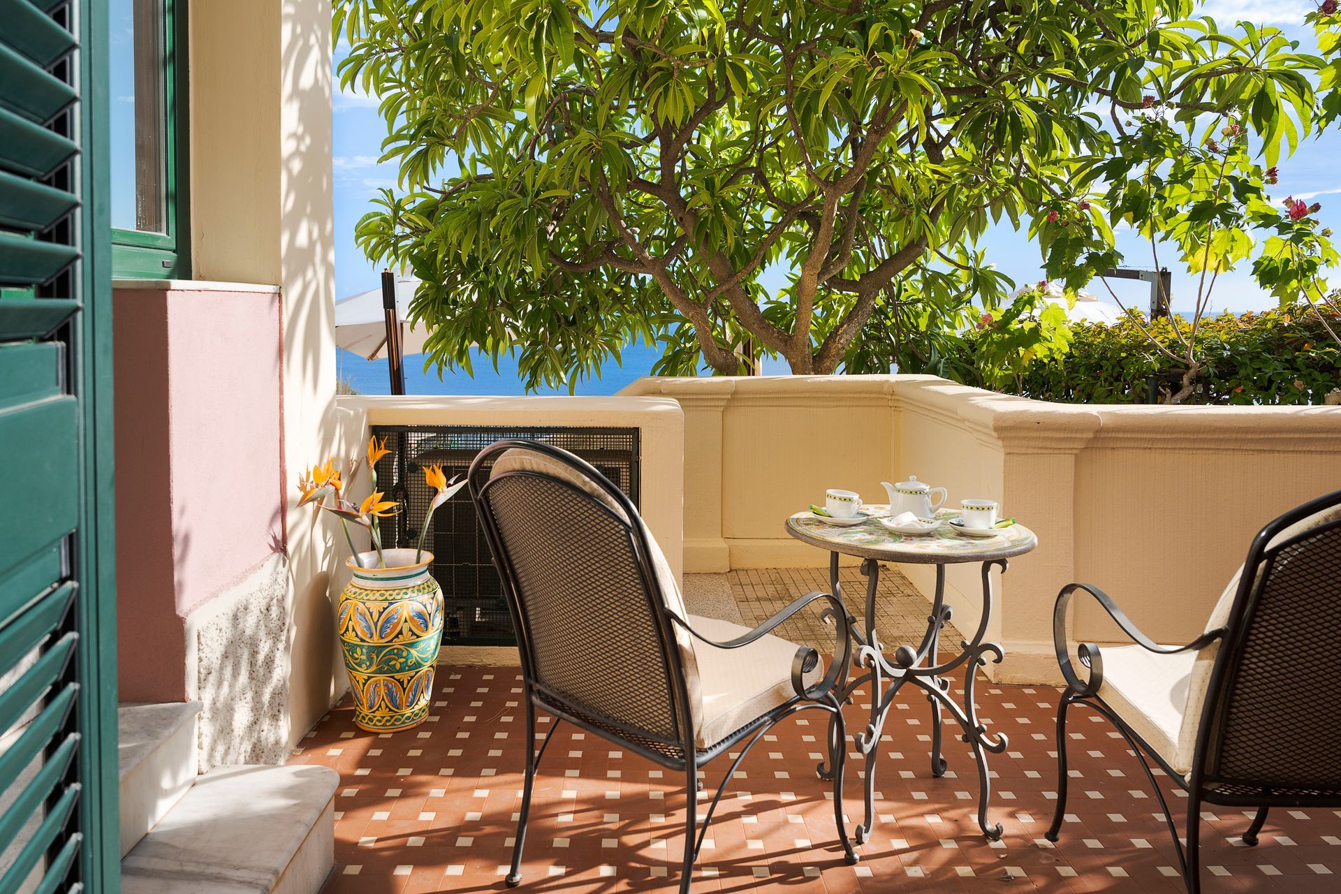 Sunlit patio with wicker chairs, small table, and potted plant beside a house and leafy tree