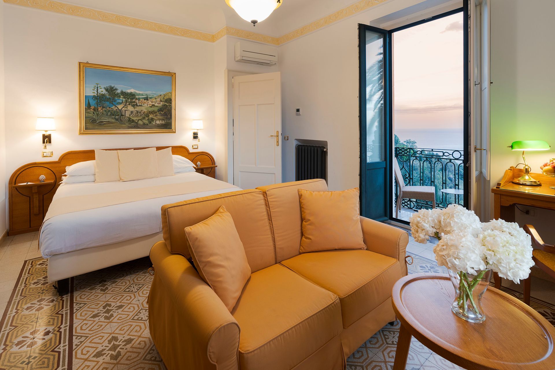 Bright hotel room with bed, tan sofa, and open balcony doors facing the sea