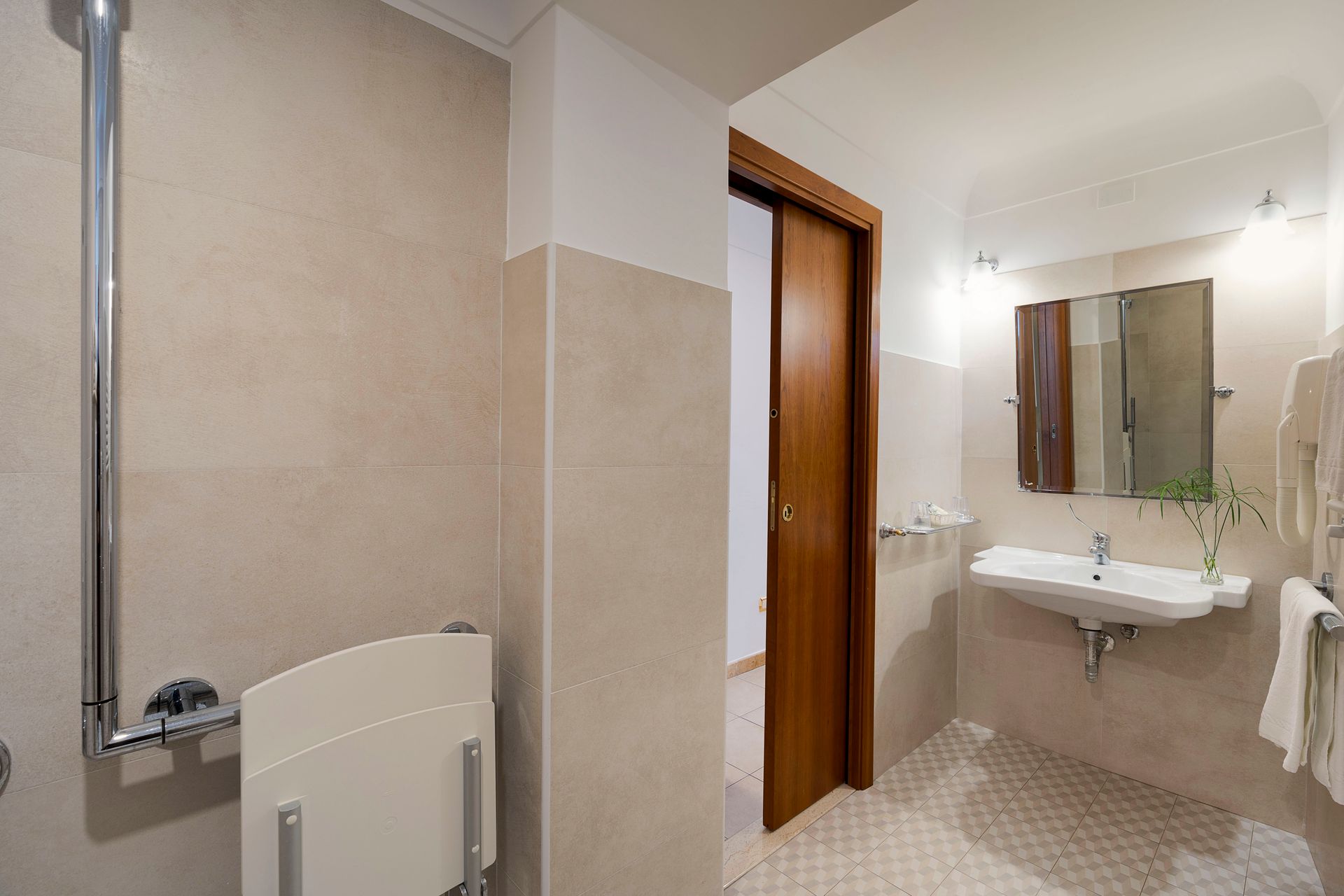 Modern bathroom with sink, mirror, toilet, beige tiles, and wood door, seen from the shower area