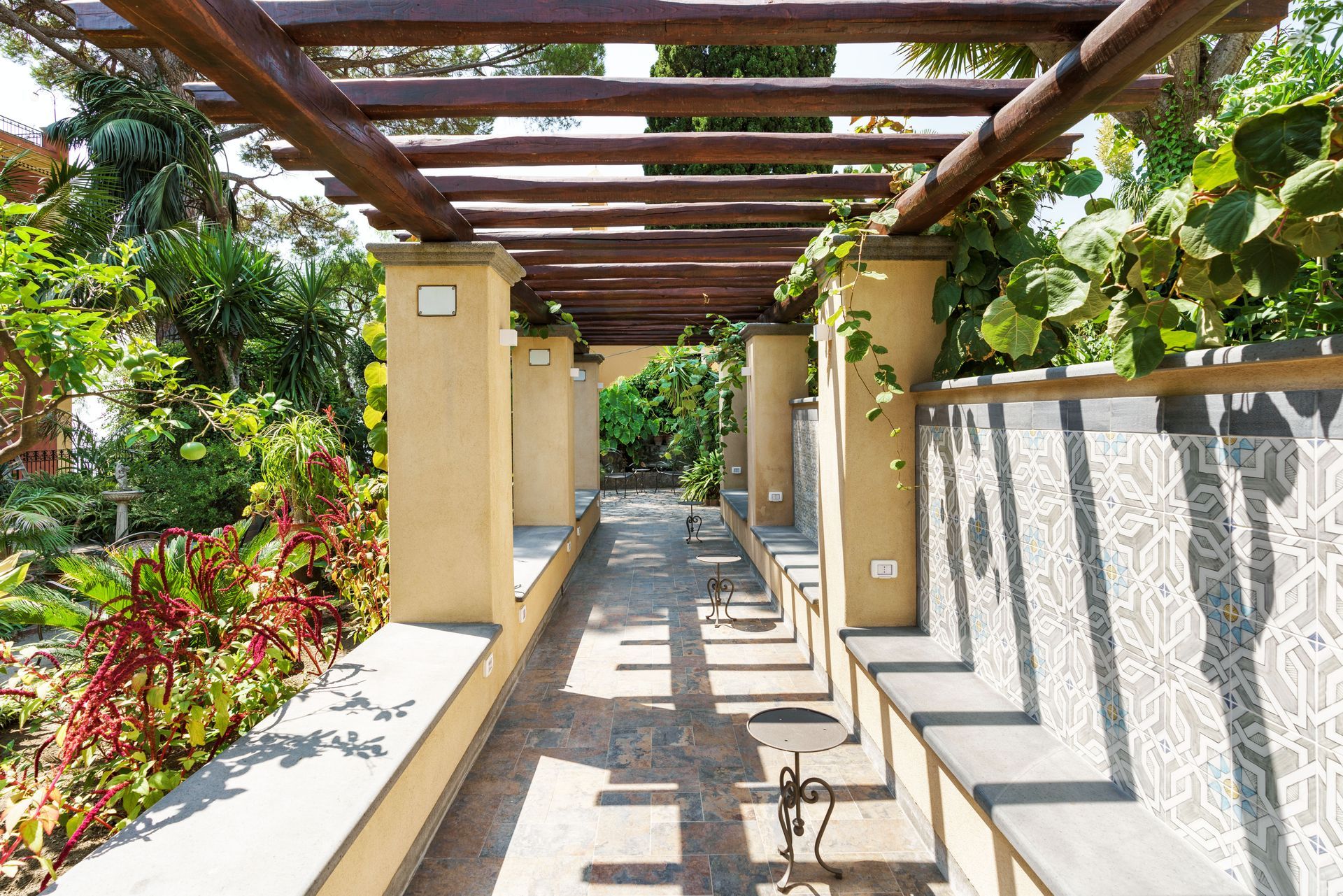 Sunlit outdoor walkway with wooden pergola, stone path, and lush greenery along the sides