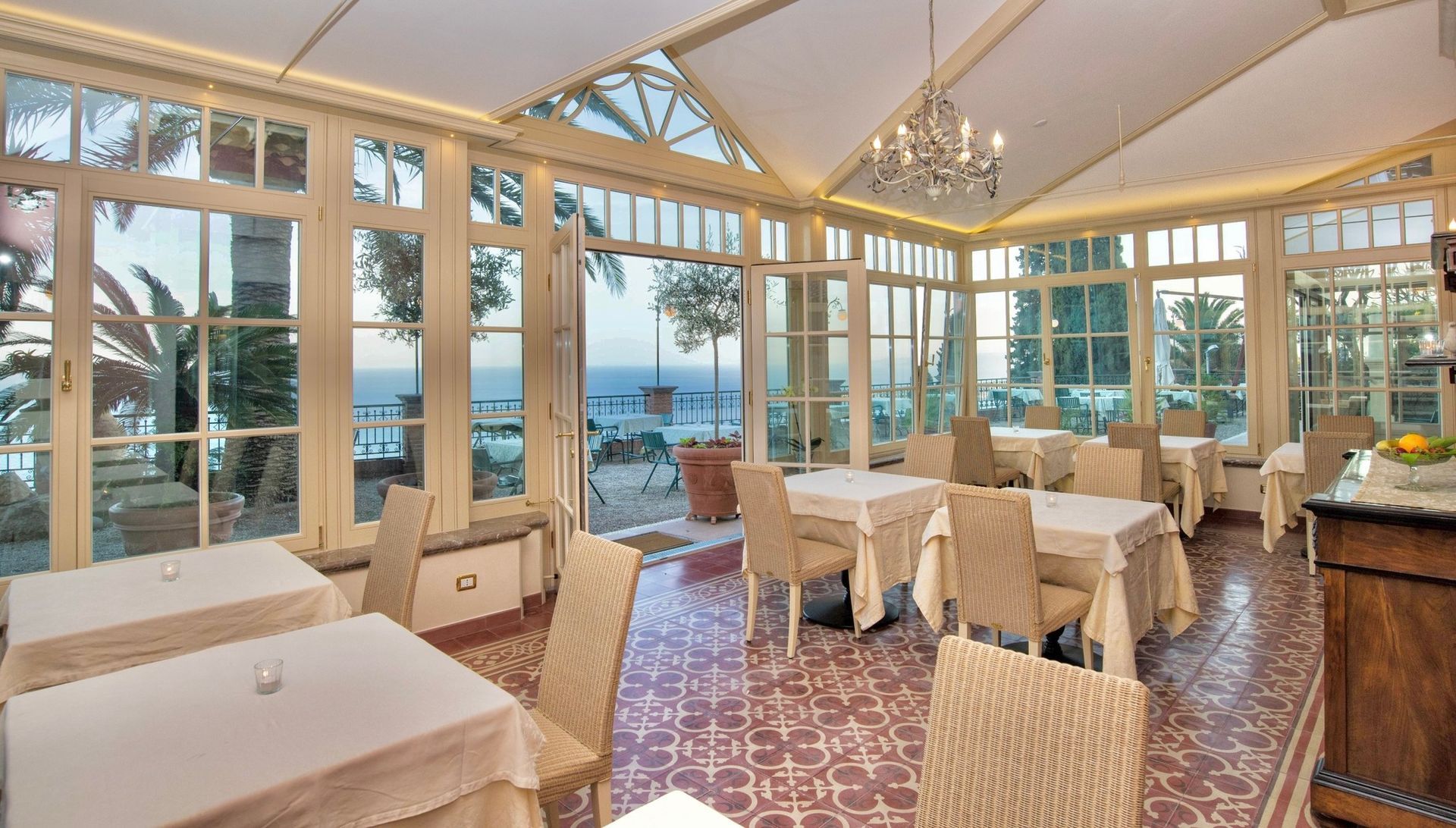 Bright dining room with large windows, white tablecloths, wicker chairs, and a view of the water