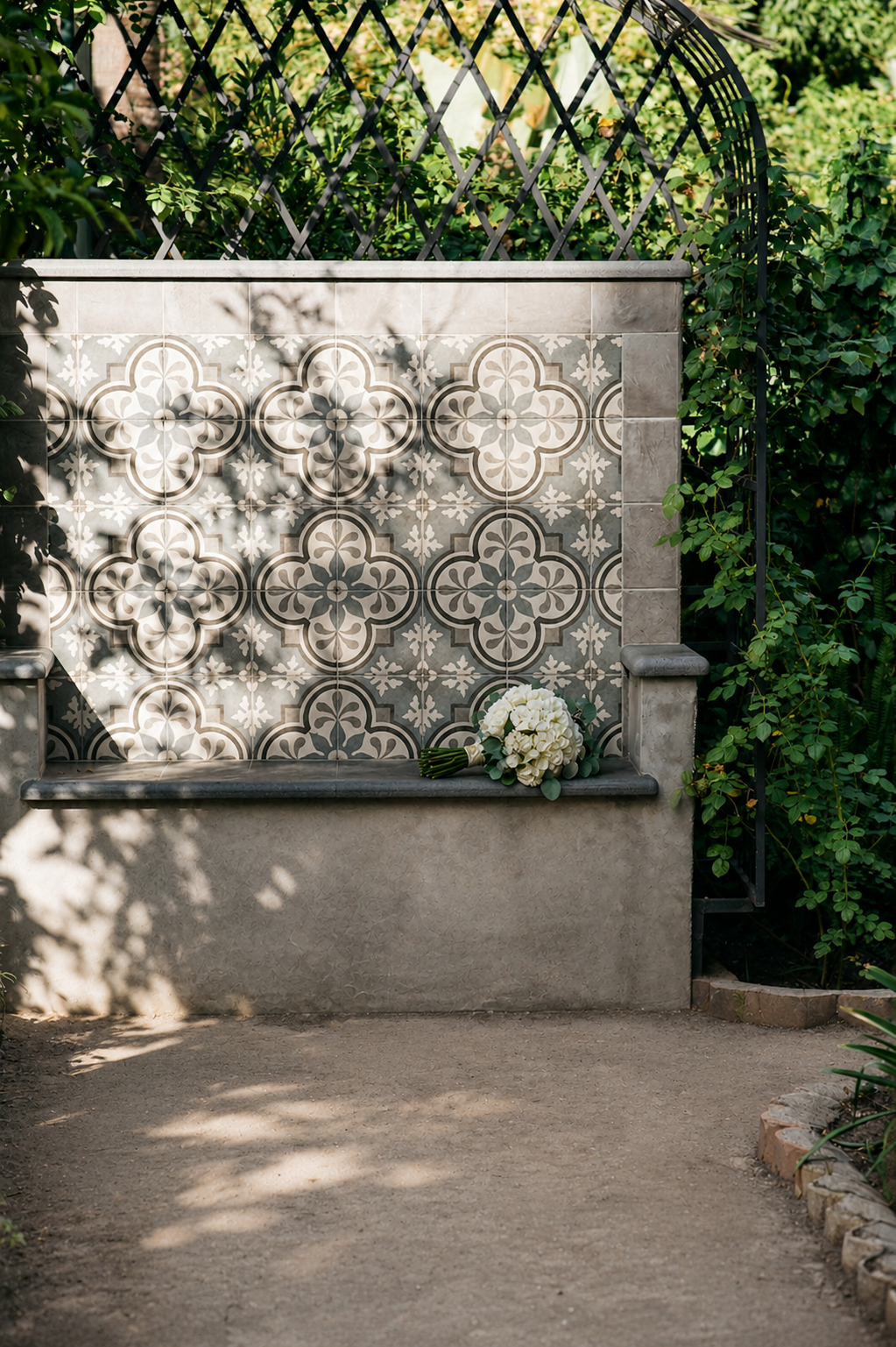 Tiled memorial wall with flowers beside a sunlit garden path and greenery
