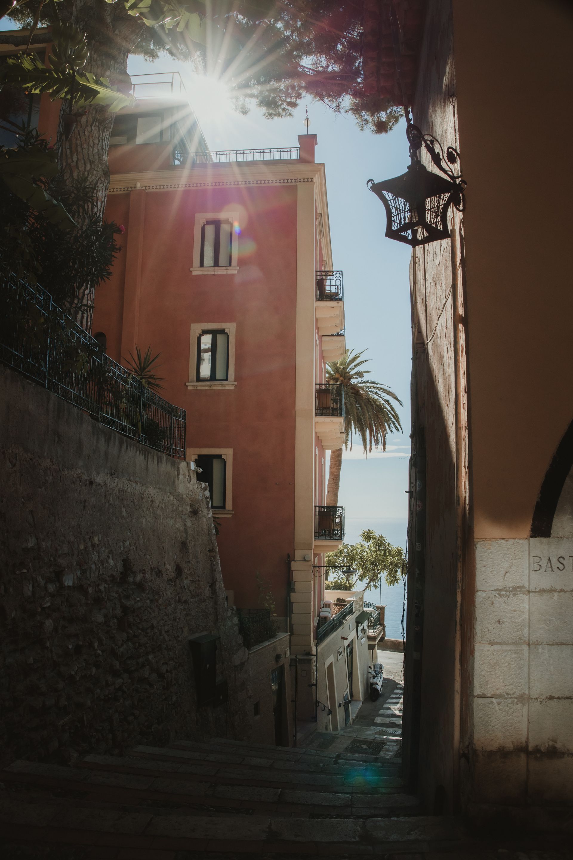 Narrow sunlit alley between tall buildings, with palm trees and bright blue sky visible at the end.