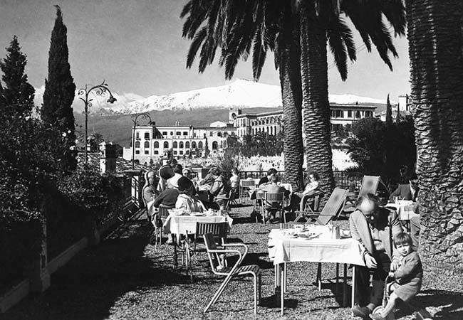 Outdoor café with seated people under palm trees, mountains and a building in the background