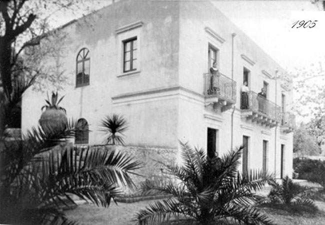 Black-and-white photo of a two-story stucco house with balcony and palm plants in front