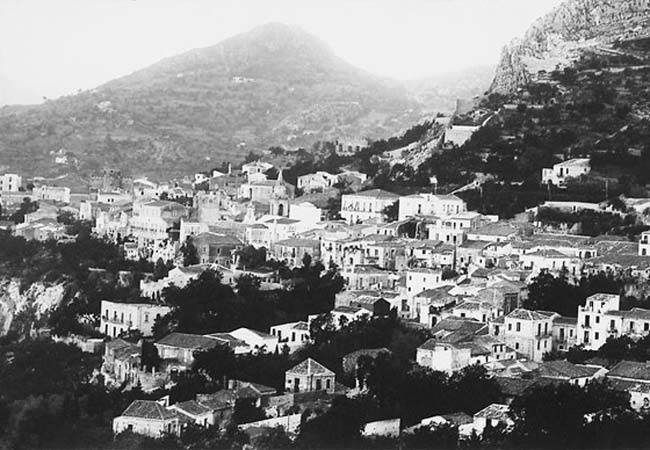 Black-and-white view of a hillside town with clustered white buildings and mountains in the background
