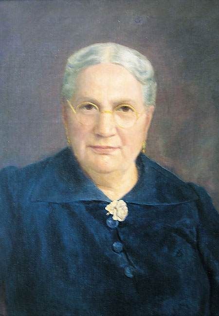 Studio portrait of an older woman in a dark blue dress with a white brooch, facing forward against a gray background