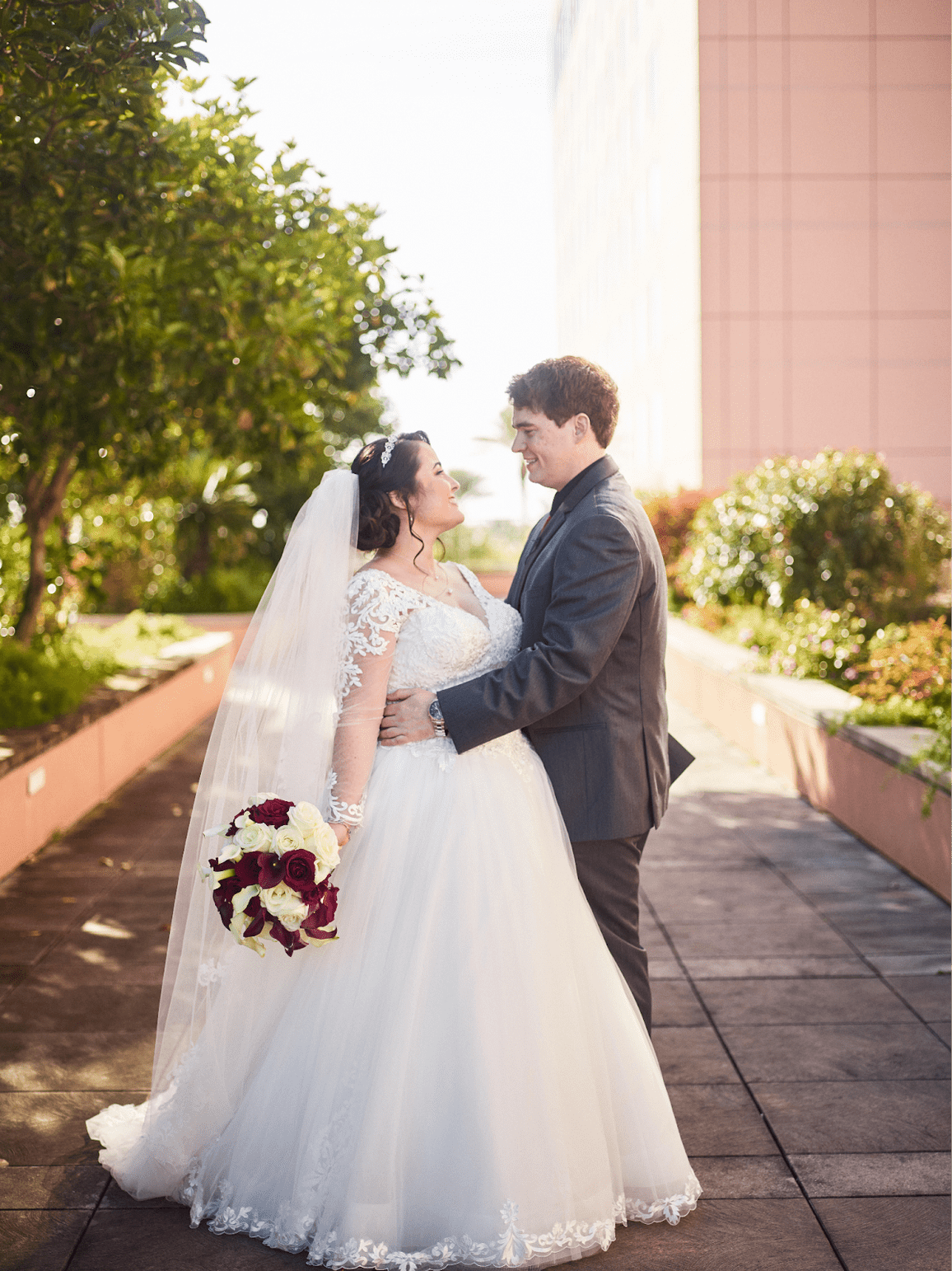 Bride and groom embracing, looking at each other. She wears a white dress, holding a bouquet. He wears a suit. Outdoors.