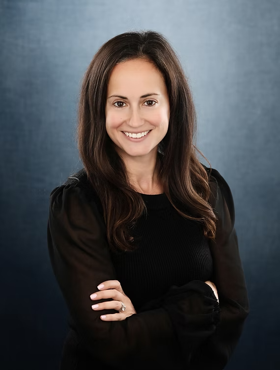 Woman with long brown hair smiles, arms crossed, wearing a black top against a blurred blue background.