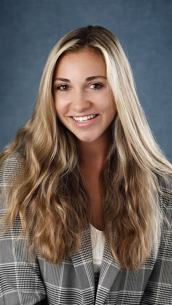 Woman with long blonde hair smiles, wearing a plaid blazer, against a blue background.