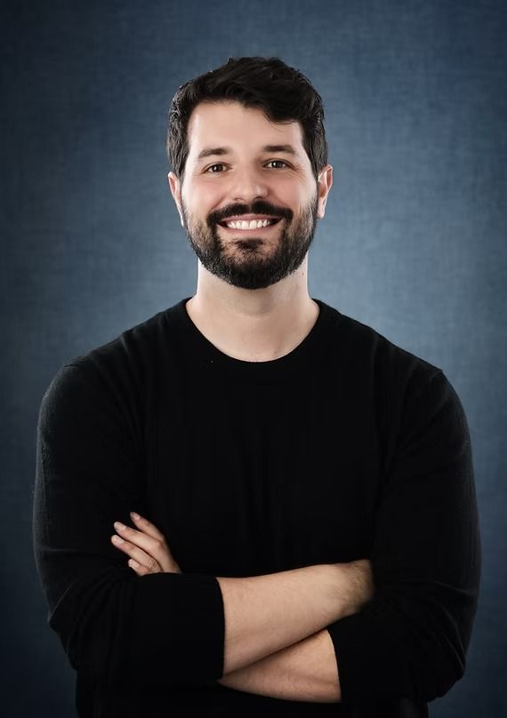 Man with dark hair and beard, arms crossed, smiling, wearing a black long-sleeve shirt, against a blue background.