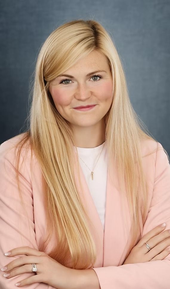 Woman with blonde hair, arms crossed, wearing a pink blazer over a white shirt, smiling.