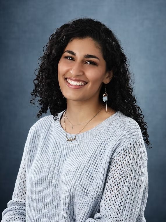 Woman with curly hair smiles, wearing a light blue sweater and a necklace, against a blue background.