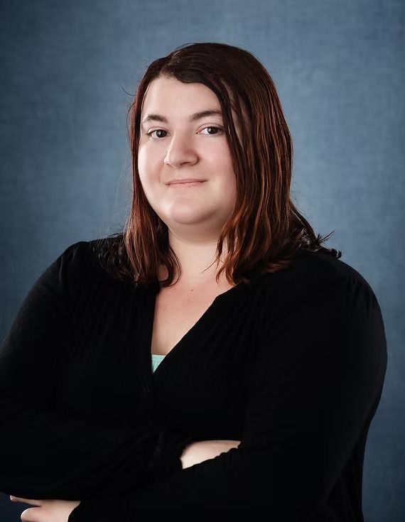 Woman with brown hair and arms crossed, wearing a black top against a blue background, smiling.