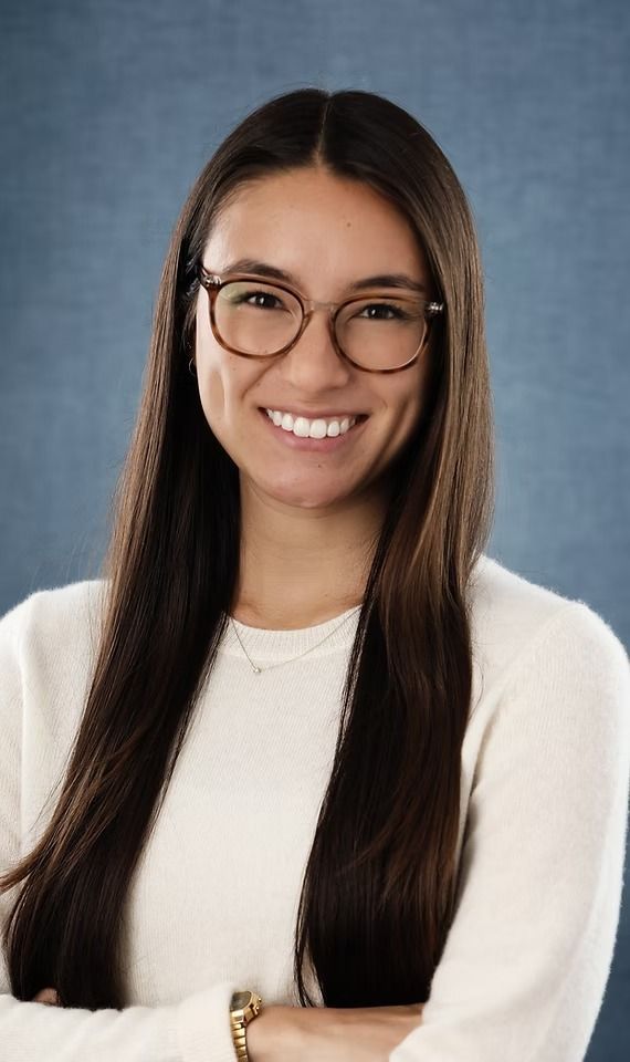 Woman with glasses, smiling with arms crossed, wearing a white sweater against a blue background.