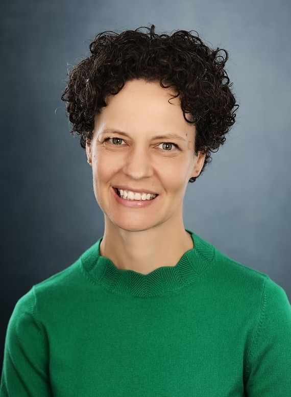 Woman with short curly hair smiles, wearing a green sweater against a dark blue background.