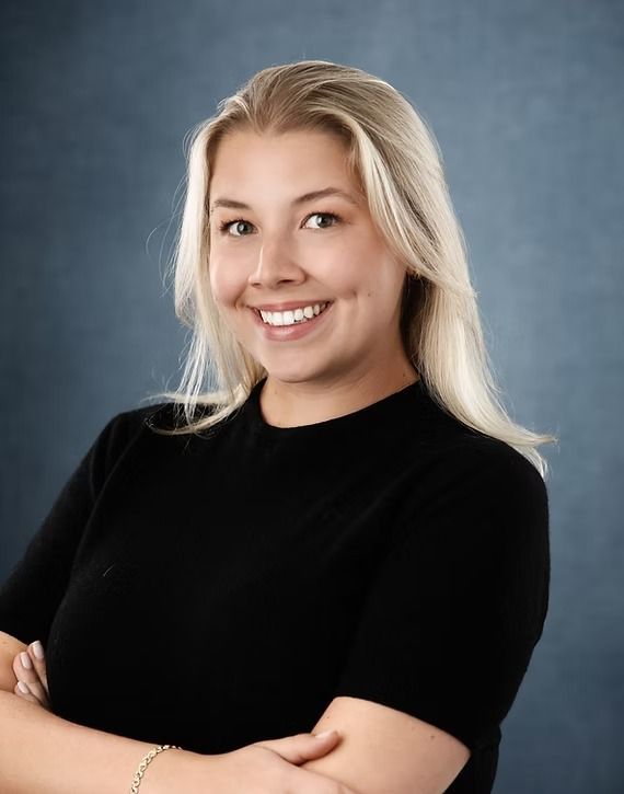 Blonde woman smiles, arms crossed, wearing a black top against a blue-gray backdrop.