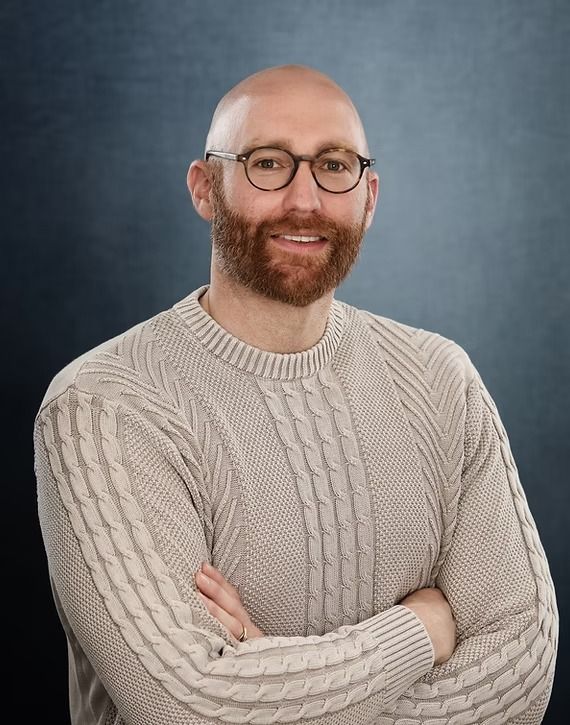 Man with glasses, bald head, and beard; arms crossed, smiles at camera. Beige sweater, blue background.