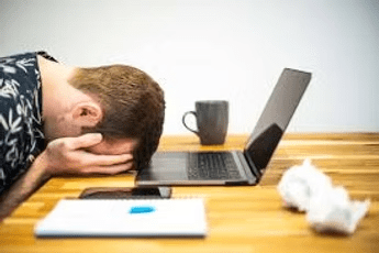 Man with head down on desk in front of laptop, appearing stressed. Mug, tissues, and notebook visible.
